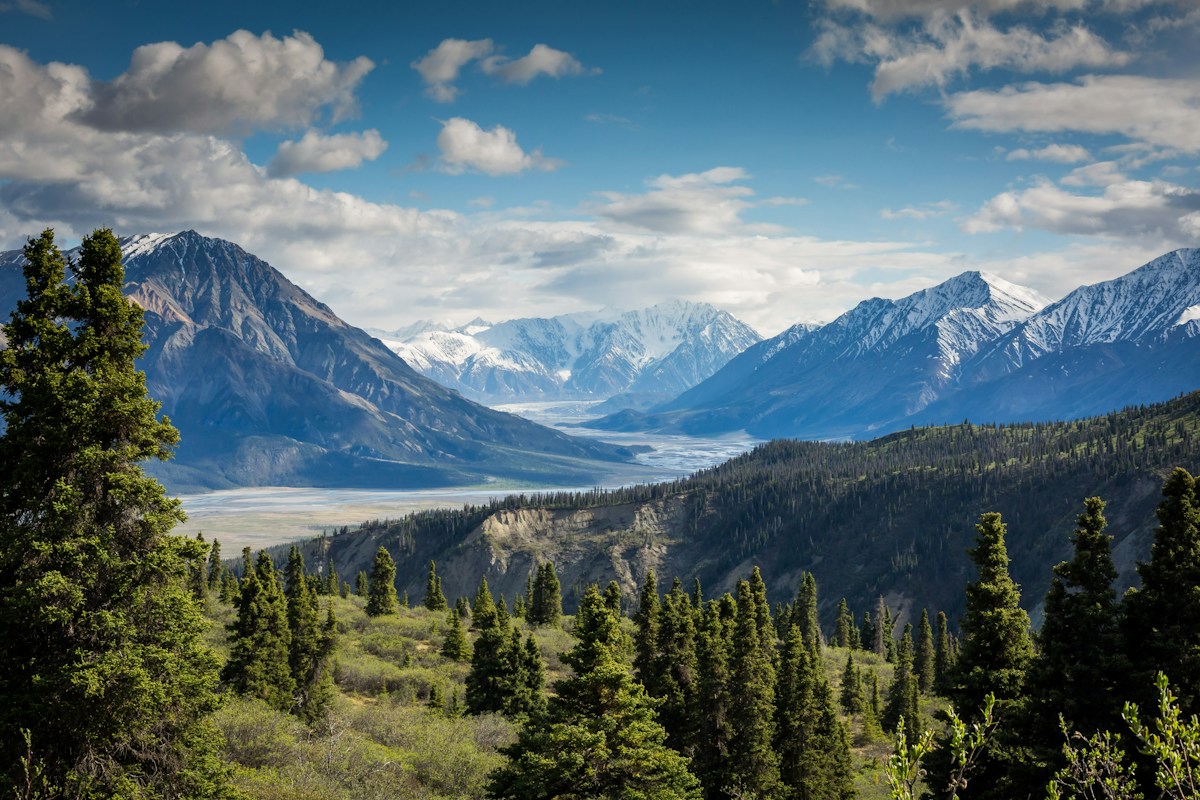 Snow-covered mountain peaks above a forested valley beneath a blue sky.